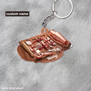 Pode incluir: Um chaveiro de metal cor ouro rosa com o nome personalizado "Henry" nele. O chaveiro tem a forma de uma bolsa de couro enrolada com um conjunto de ferramentas de coquetel dentro.