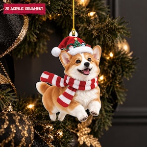 Puede incluir: Un adorno de acrílico 2D que muestra un perro Corgi de color canela y blanco con un gorro de Papá Noel rojo y blanco, un pequeño árbol de Navidad y una bufanda a rayas rojas y blancas. El adorno cuelga de un cordón dorado.