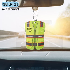 May include: A yellow safety vest with reflective stripes and black accents. The vest has a name tag that says "Jannee" and the words "Crossing Guard" printed on it. The vest is hanging from a silver chain.