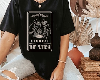 Könnte beinhalten: Schwarzes T-Shirt mit einem Grafikdruck einer Tarotkarte, die eine Hand mit einer Kristallkugel zeigt, darunter der Text "The Witch".