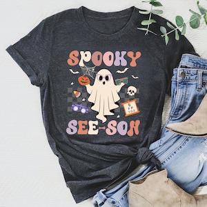 Może przedstawiać: Szary t-shirt z nadrukiem ducha w okularach i napisem "Spooky See-Son" w kolorowych, retro literach. Duch trzyma dynię, a wokół niego znajdują się inne grafiki związane z Halloween.