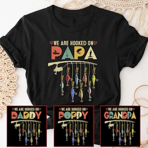 May include: A black t-shirt with a vintage-style graphic of a fishing rod with lures. The text "We are hooked on Papa" is above the graphic. Three smaller versions of the graphic are below, with the text "We are hooked on Daddy", "We are hooked on Poppy", and "We are hooked on Grandpa".