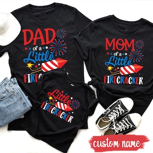 Puede incluir: Tres camisetas negras con temática del 4 de julio. Las camisetas presentan un diseño de fuegos artificiales rojo, blanco y azul con el texto "Papá de un pequeño petardo", "Mamá de un pequeño petardo" y "Pequeño petardo".