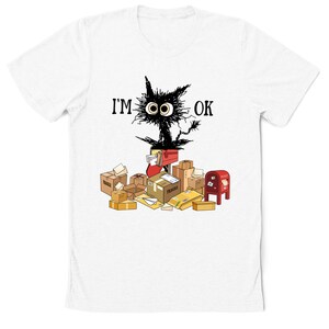 May include: White t-shirt with a black cartoon cat surrounded by boxes and mail. The cat has large eyes and the text "I'M OK" above it.
