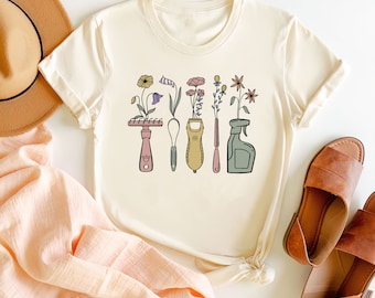 Może przedstawiać: Biały t-shirt z grafiką narzędzi ogrodniczych, z których rosną kwiaty. Narzędzia obejmują grabie, łopatę, ręczny kultywator, sekator i butelkę z rozpylaczem. Kwiaty są w odcieniach różowego, fioletowego i żółtego.
