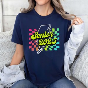 May include: A navy blue t-shirt with a rainbow checkered pattern and a lightning bolt graphic. The text "Senior 2025" is written in yellow on the lightning bolt.