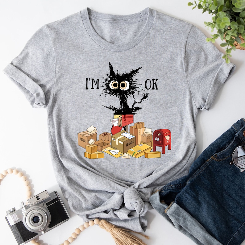 May include: A gray t-shirt with a black cat illustration and the text "I'M OK". The cat is surrounded by boxes and envelopes, suggesting a busy mailroom or delivery service.