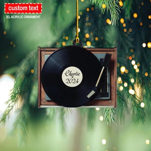 Puede incluir: Un adorno de tocadiscos marrón y blanco con un disco de vinilo negro. El disco tiene una etiqueta blanca con el texto "Charlie 2024".