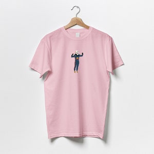 以下が含まれることがあります： 青いジャンプスーツを着た人物の漫画イラストが描かれたピンクのTシャツです。人物は両手を上げています。
