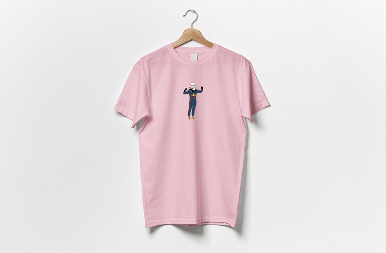以下が含まれることがあります： 青いジャンプスーツを着た人物の漫画イラストが描かれたピンクのTシャツです。人物は両手を上げています。
