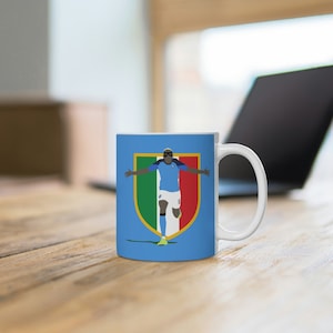 Pode incluir: Uma caneca de cerâmica branca com fundo azul e um gráfico de um jogador de futebol com uma camisa azul com a bandeira italiana em um escudo. O jogador está comemorando um gol com os braços levantados.