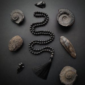 Könnte beinhalten: Eine schwarze Lavaperlenkette mit schwarzem Quastenanhänger, angeordnet mit verschiedenen dunklen Steinen und Fossilien auf schwarzem Untergrund. Die Kette ist der Mittelpunkt, umgeben von natürlichen Elementen.