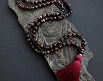 May include: A beaded necklace with deep red gemstones and silver-colored spacers. The necklace has a matching red tassel and rests on a dark gray, carved stone with a face design. The background is a solid black.