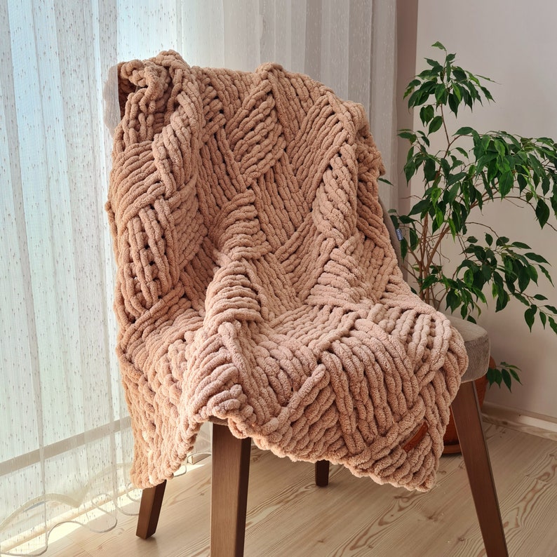 May include: A soft, beige knitted throw blanket with a woven pattern draped over a wooden chair.