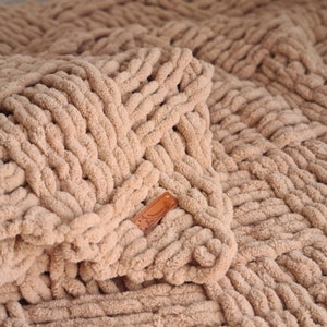 May include: A close-up of a beige knitted throw blanket with a woven texture. The blanket has a brown leather tag with the word "Handmade" on it.