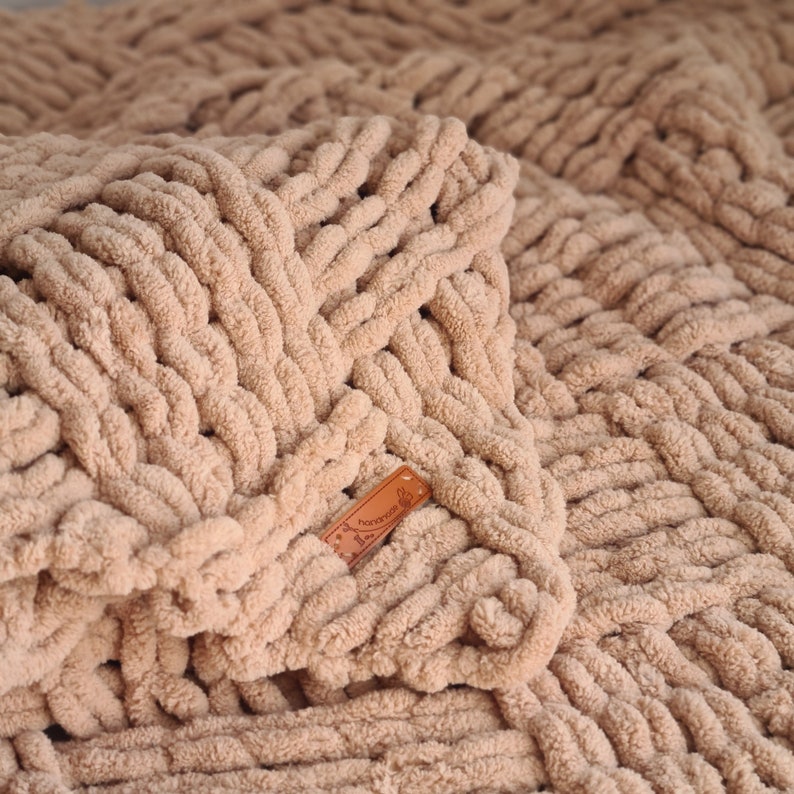 May include: A close-up of a beige knitted throw blanket with a woven texture. The blanket has a brown leather tag with the word "Handmade" on it.