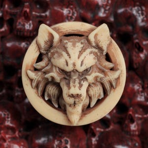 May include: A beige wolf head pendant with a circular frame, set against a backdrop of numerous red skulls. The wolf's face is detailed with sharp features. The skulls have a dark, aged appearance.