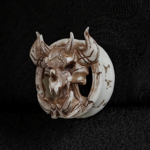 May include: An off-white and brown ear gauge plug with a detailed demonic skull design. The skull has horns and sharp teeth, set within a circular frame. The plug is likely made of a durable material, suitable for body modification.