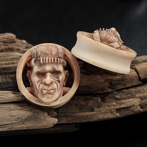 以下が含まれることがあります： ベージュ色の2つの耳栓。フランケンシュタインのモンスターのデザインが彫られています。1つはモンスターの顔を円形のフレームで、もう1つは頭頂部を表示しています。耳栓は風化した木材の上に置かれています。