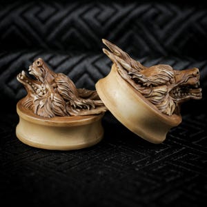 May include: A pair of beige ear gauges, each featuring a carved wolf head. The wolf heads are intricately detailed, with open mouths and visible teeth. The gauges are set against a dark, textured backdrop, highlighting the carvings.