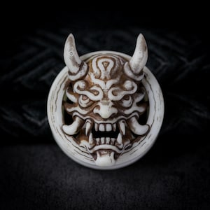 May include: A detailed, off-white and brown, circular sculpture of a Japanese Oni mask. The mask features prominent horns, sharp teeth, and intricate facial details. The sculpture is set against a dark, textured background.