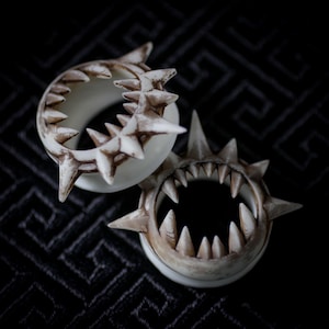 Puede incluir: Dos expansores de oreja de color marfil con diseño de mandíbula de tiburón. Cada expansor presenta dientes afilados y puntiagudos y púas alrededor del borde exterior, con acentos marrones. Los expansores están sobre un fondo oscuro estampado.