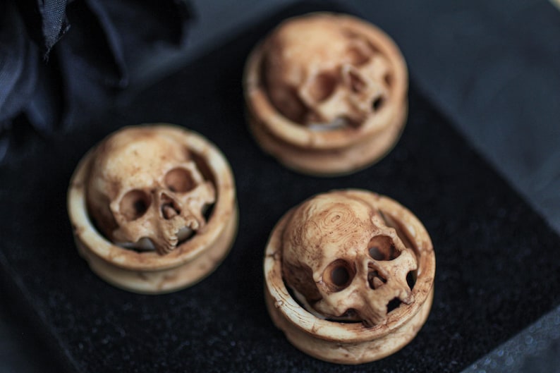 May include: Three round, wooden ear gauges with a skull design. The skulls are carved in detail and have a light brown color.
