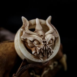 May include: A detailed, off-white sculpture of a dragon's head with horns, sharp teeth, and a circular frame. The sculpture is set against a dark background, highlighting the intricate details and textures of the artwork. The dragon's face is centered.