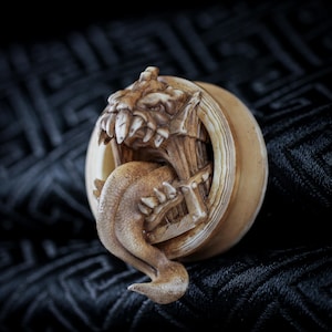 Pode incluir: Escultura detalhada, de cor bege, de uma criatura mítica emergindo de uma forma circular. A criatura tem dentes afiados e uma língua longa e texturizada. A escultura está sobre um fundo escuro e padronizado.