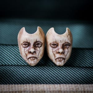 May include: Two carved, off-white masks with detailed facial features and curved horns. The masks have etched lines and a slightly aged appearance, set against a textured green background. The masks are symmetrical and appear to be decorative or artistic.