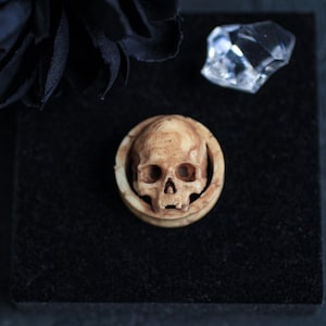 May include: A close-up of a skull-shaped ear gauge made of bone-colored material. The gauge is on a black velvet surface.