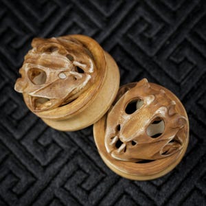 Könnte beinhalten: Zwei Ohrstöpsel aus Holz mit einem geschnitzten Design eines stilisierten Schädels. Die Stöpsel sind hellbraun mit einem helleren, fast weißen Innenraum. Der Hintergrund ist ein dunkler, strukturierter Stoff.