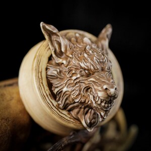 Könnte beinhalten: Eine detaillierte, geschnitzte Holzskulptur eines Wolfskopfes, der aus einem kreisförmigen Rahmen hervortritt. Der Wolf hat einen wilden Ausdruck, mit sichtbaren Zähnen und strukturierter Fell. Das Stück ist in Braun- und Beigetönen vor dunklem Hintergrund.