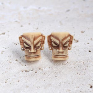 Op de afbeelding: Twee kleine, beige tiki-stijl sculpturen met gesneden details. Elk heeft een streng gezicht met schuine ogen, een brede mond en gedetailleerde oorvormen. De sculpturen worden gepresenteerd op een getextureerd, gebroken wit oppervlak.