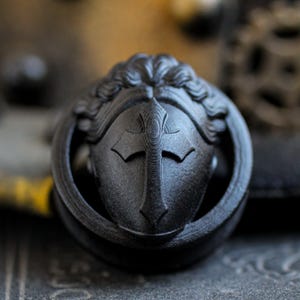 May include: Black ear gauge plug with a detailed design of a face with a cross on the forehead.