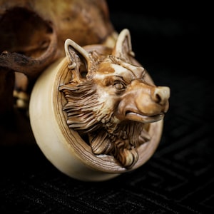 Könnte beinhalten: Eine detaillierte, geschnitzte Holzskulptur eines Wolfskopfes. Der Wolf ist mit aufwendigem Fell und Gesichtszügen dargestellt, eingebettet in einen kreisförmigen Rahmen. Die Skulptur ist hauptsächlich hellbraun mit dunkelbraunen Akzenten, die die Holzmaserung zeigen.