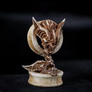 May include: A detailed, off-white sculpture of a demonic creature with sharp teeth and horns. The sculpture features a circular element with a similar face and is set on a round base. The artwork has a textured appearance and is set against a black background.