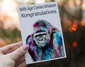 May include: Greeting card with a watercolor painting of a gorilla. The gorilla is depicted with vibrant splashes of blue, pink, and purple. The card has the text "With Age Comes Wisdom Kongratulations" in black font.