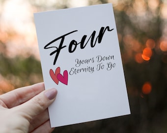 Peut inclure: Une carte blanche avec du texte noir qui dit "Four Years Down Eternity To Go". Deux cœurs roses sont dessinés sur la carte.