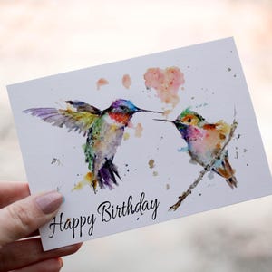Puede incluir: Tarjeta de cumpleaños con una ilustración en acuarela de dos colibríes. Uno está en vuelo, el otro posado en una rama. La tarjeta tiene la inscripción "Happy Birthday" en escritura cursiva.