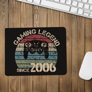 Peut inclure: Tapis de souris noir avec le texte "GAMING LEGEND SINCE 2006" et un graphisme de manette. Le design présente un style rétro avec un motif circulaire de rayures horizontales dans des tons de bleu, vert et marron.
