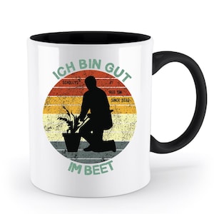 Puede incluir: Taza de cerámica blanca con interior y asa negros. La taza presenta un diseño desgastado con un jardinero arrodillado, una planta y el texto "ICH BIN GUT IM BEET" en verde y rojo. El diseño está sobre un fondo de puesta de sol.