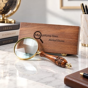 May include: A magnifying glass with a gold-toned rim and a carved wooden handle featuring floral details. It is positioned in front of a rectangular wooden storage box engraved with the text "Magnifying Glass" and "Michael Owens".
