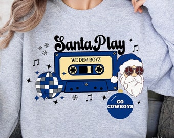 Puede incluir: Sudadera gris con un gráfico de cinta de casete azul y blanco, una bola de discoteca, un Papá Noel con gafas de sol y un adorno azul que dice "Go Cowboys". El texto en la cinta de casete dice "We Dem Boyz" y el texto sobre la cinta de casete dice "Santa Play".