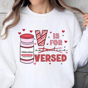 May include: White sweatshirt with a red and white striped letter V, a red heart, and the text "V is for Valentine Versed". A small pink bottle with the text "Midazolam Injection, USP" is next to the V.