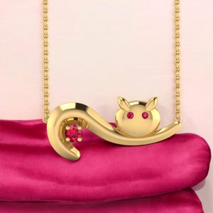 Puede incluir: Un collar con colgante dorado con un diseño de gato estilizado. El gato tiene ojos de color rubí y una piedra preciosa de rubí. El collar se muestra sobre una superficie rosa.