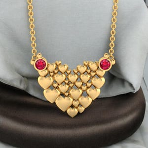 May include: A gold-colored necklace featuring a heart-shaped pendant. The pendant is composed of numerous small hearts and two red gemstones. The necklace has a delicate chain and is displayed on a neutral background.