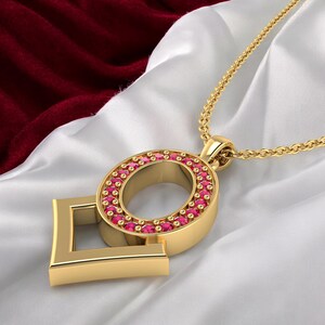 May include: A gold pendant necklace with a letter "O" design, encircled by small, red gemstones. The pendant is attached to a fine gold chain. The background is white satin with a burgundy velvet accent.
