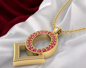 May include: A gold-colored pendant necklace featuring a stylized letter "O" with a ring of small, red gemstones. The pendant is connected to a delicate gold chain. The background is a white satin fabric with a burgundy velvet accent.