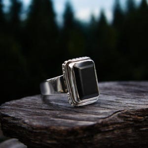 Può includere: Un anello d'argento con una grande pietra nera rettangolare. L'anello ha un bordo dettagliato e strutturato attorno alla pietra e una fascia larga. L'anello è esposto su un pezzo di legno, con uno sfondo sfocato di alberi.